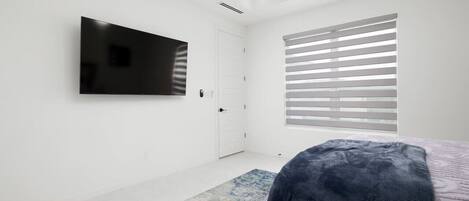 A modern bedroom with a flat-screen TV mounted on the wall, a large window with blinds, and a patterned rug on the floor.