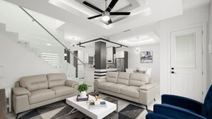 Ein modernes Wohnzimmer mit zwei beigen Sofas, einem weißen Couchtisch und einem blauen Sessel. Ein Deckenventilator und Einbaulampen sind sichtbar.