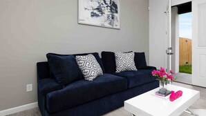 Un soggiorno moderno con un divano blu scuro, cuscini decorati, un tavolino bianco con un libro e un oggetto decorativo rosa, e un quadro appeso al muro.