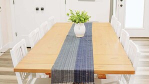 Een eettafel met witte stoelen, een blauw-grijs tafelkleed en een witte vaas met groene planten.