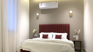 Ein Hotelzimmer mit einem Bett, weißen Vorhängen, einer Klimaanlage, zwei Wandleuchten und einer Blumenvase auf einem Nachttisch.