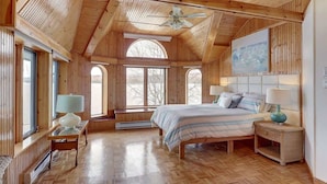 A wooden cabin bedroom with a bed, two bedside tables, a ceiling fan, and large windows.