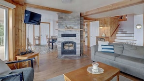 A cozy living room with a fireplace, wooden beams, a flat-screen TV, and a staircase leading to an upper level.