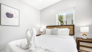 Ein Hotelzimmer mit einem großen Bett, einem Nachttisch mit Lampe, einem gerahmten abstrakten Kunstwerk und einem Fenster mit Blick auf Gebäude und Bäume.
