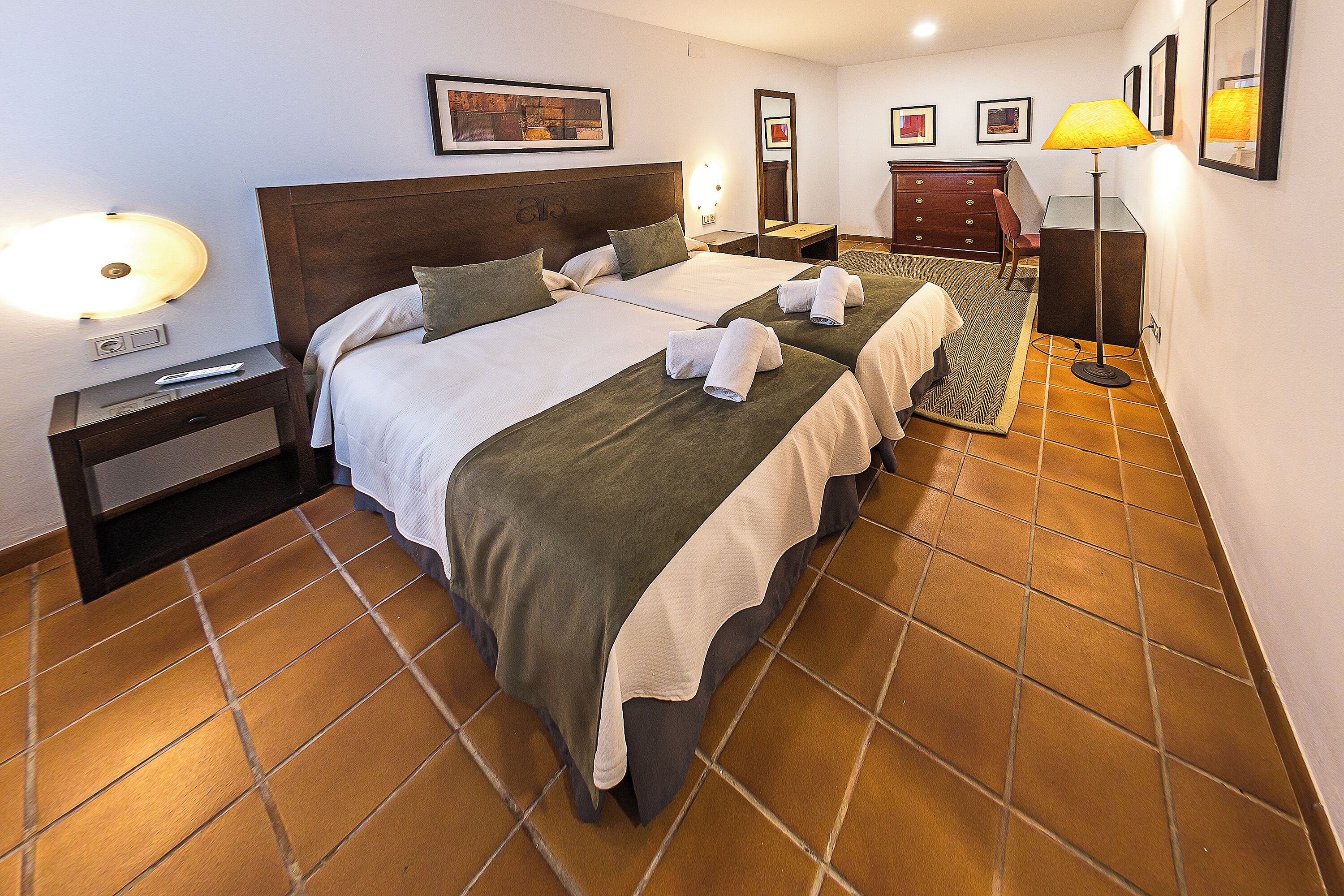 A hotel room with two beds, a nightstand, a desk, a chair, and a lamp.