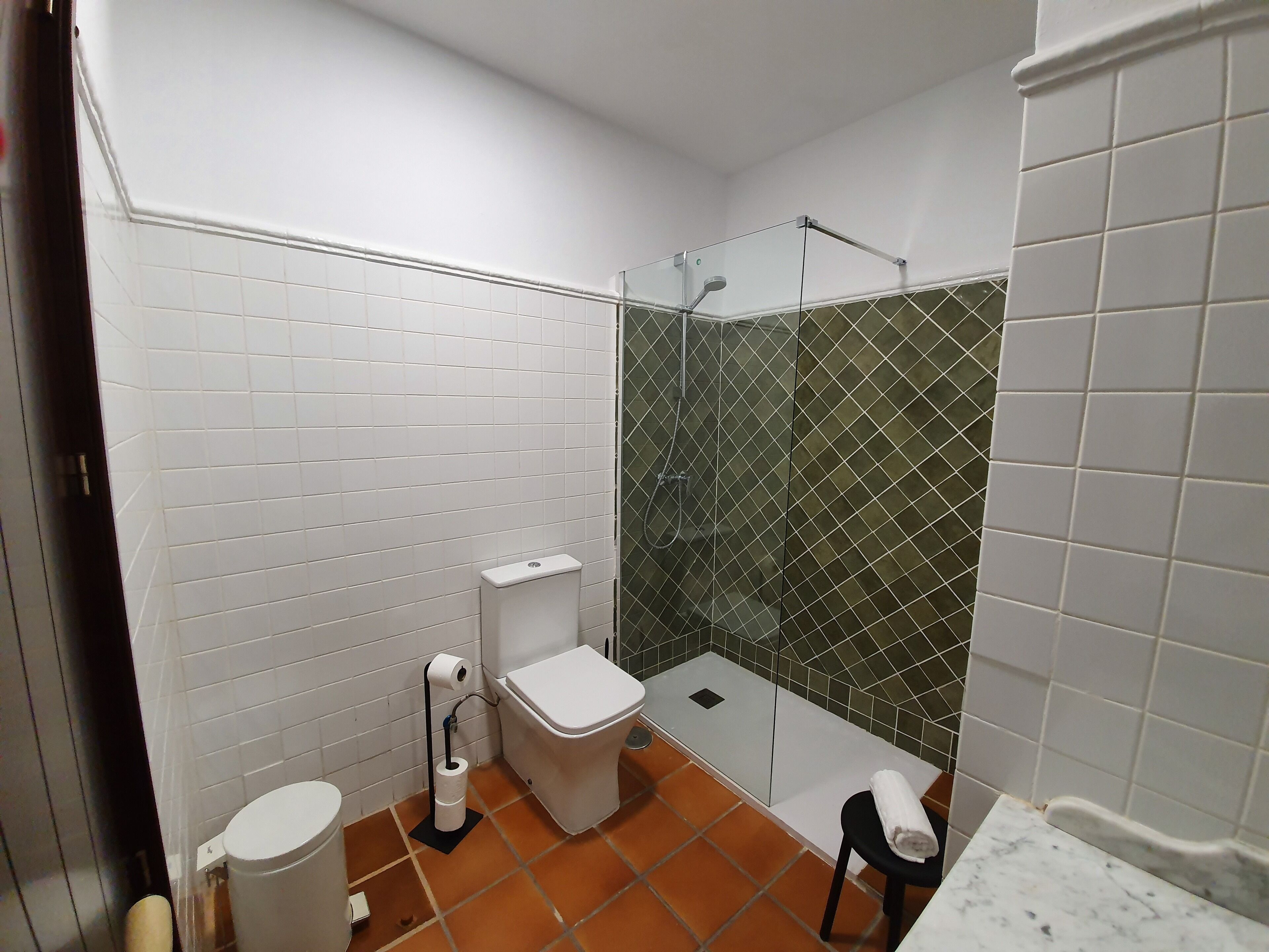 A bathroom with white tiled walls, a toilet, a shower with a glass door, and a small stool with a towel.