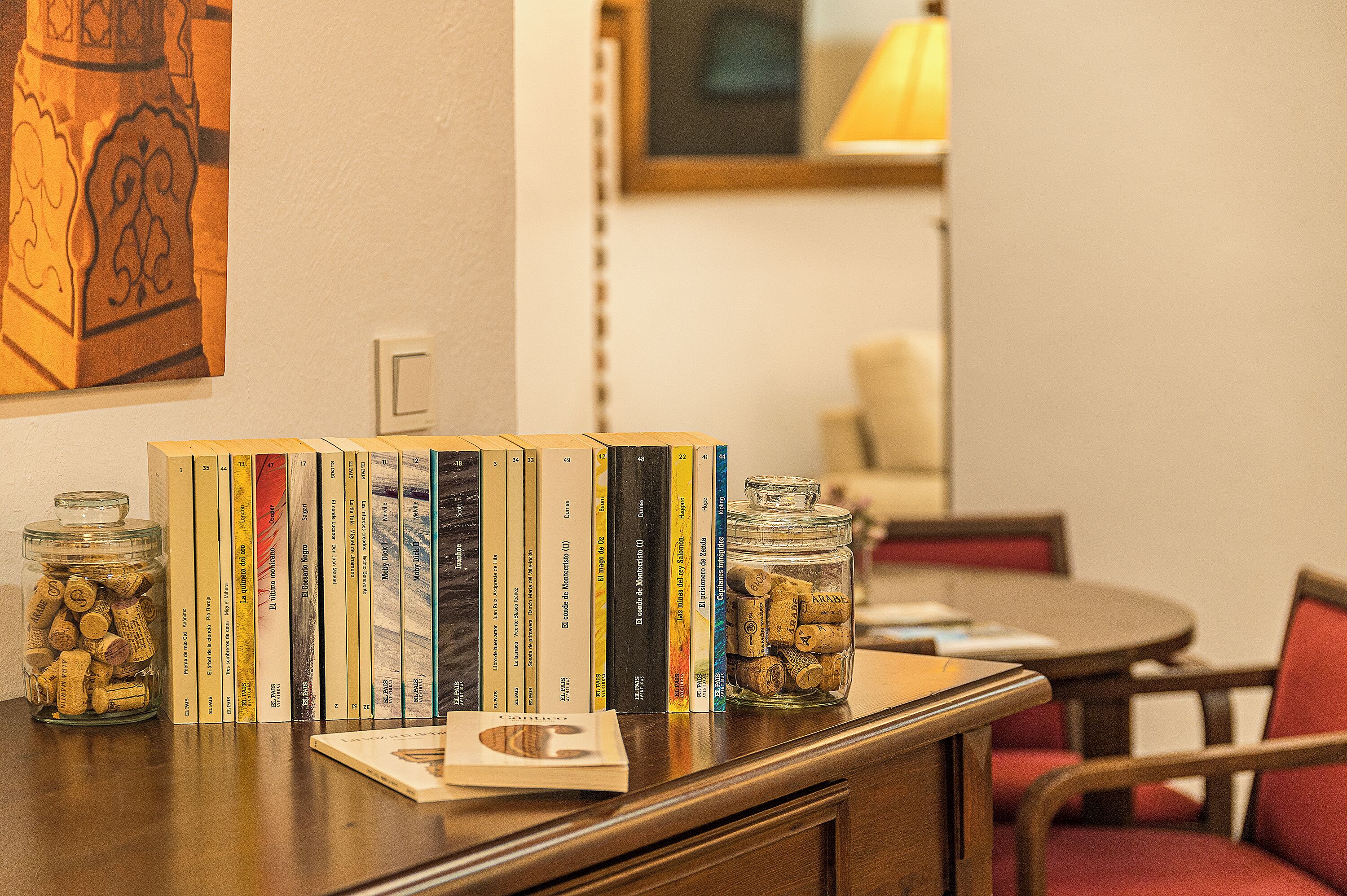 A table with books, a jar of corks, and a lamp in the background.