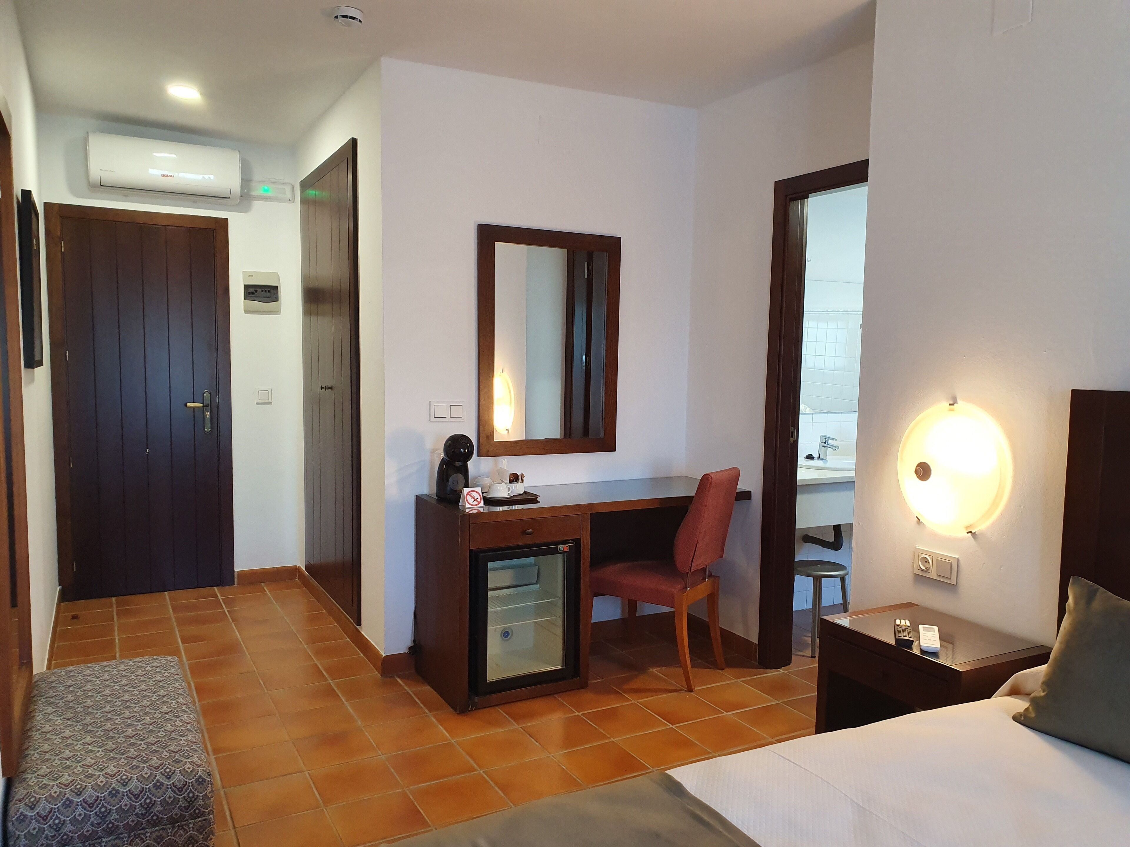 Habitación de hotel con cama, escritorio con silla, un refrigerador pequeño, espejo y mesita de noche con lámpara.