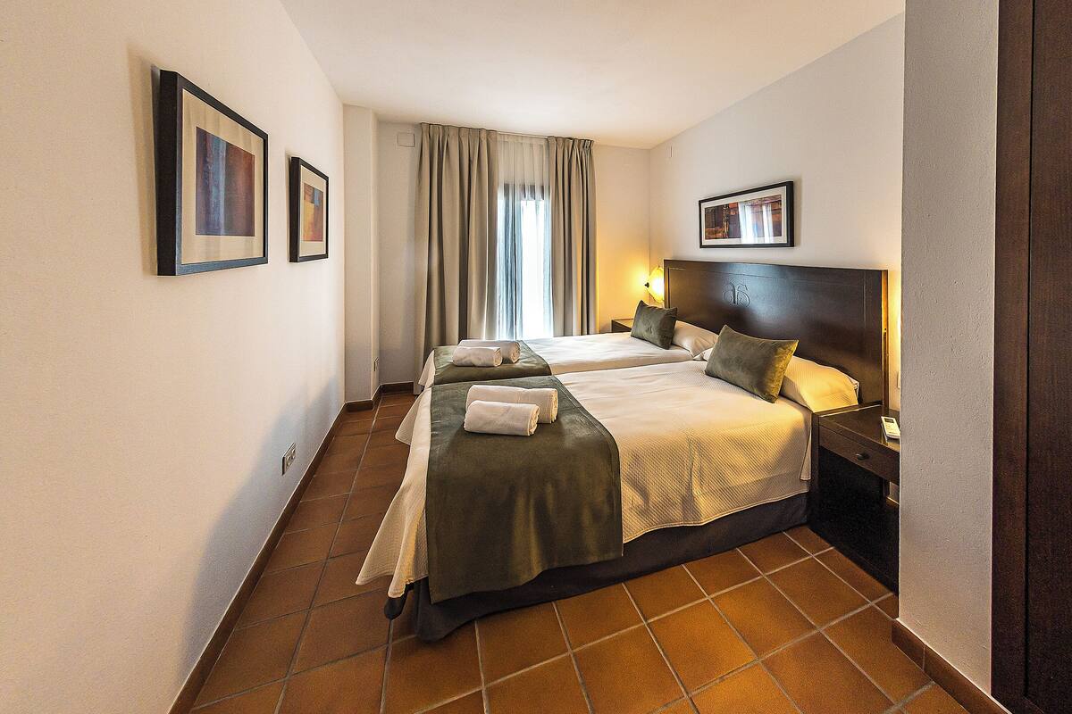 A hotel room with two beds, a large window with curtains, a bedside table with a lamp, and framed pictures on the walls.