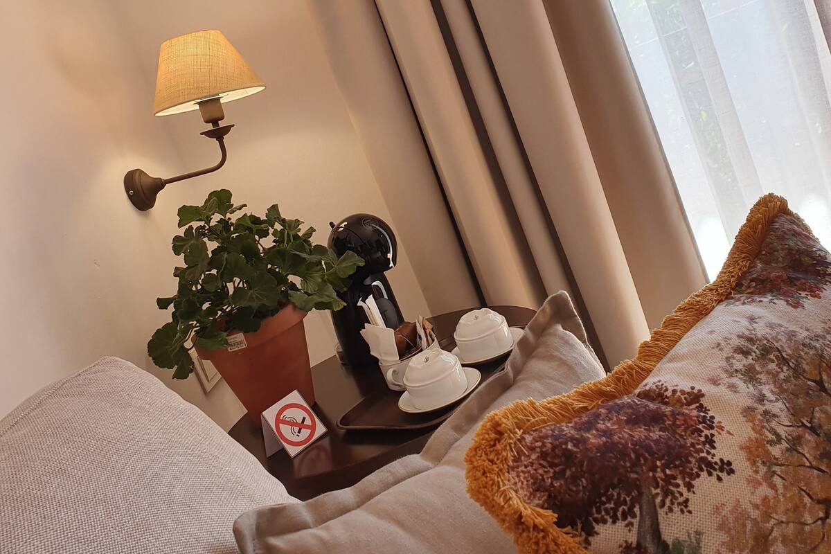A neatly arranged hotel room with a lamp, a potted plant, a coffee maker, and a table with a no-smoking sign.