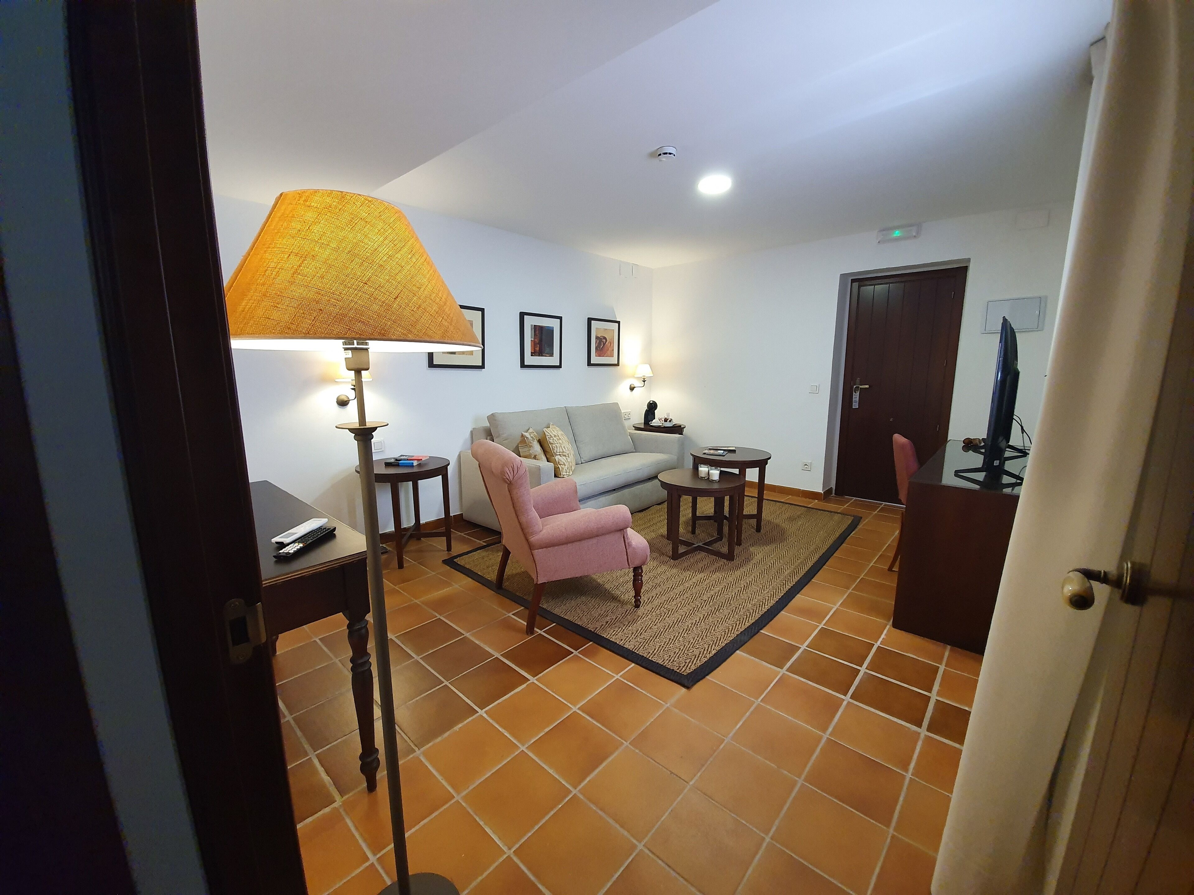 Una sala de estar con un sofá, un sillón rosa, una lámpara, una mesita y un televisor sobre un mueble bajo.