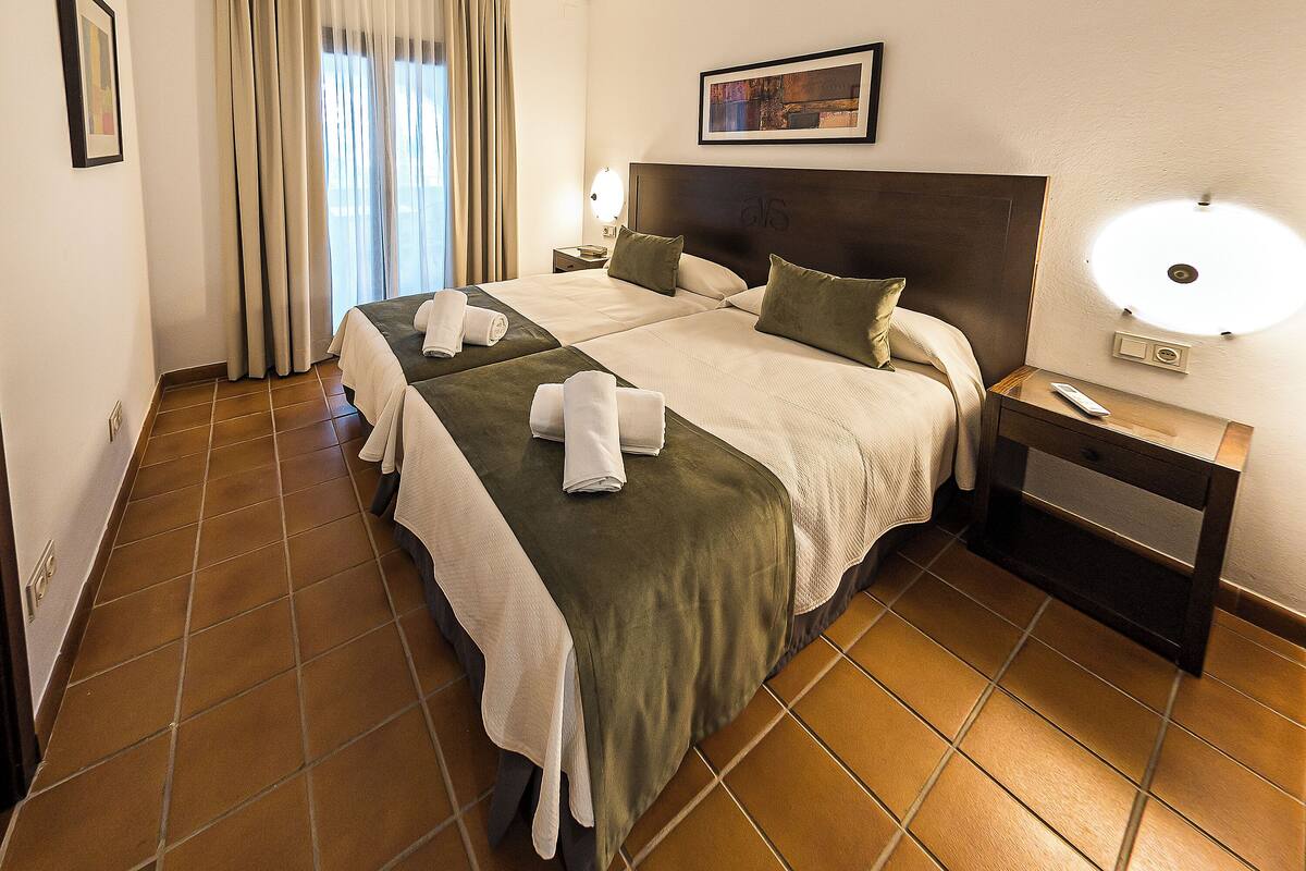 A hotel room with two beds, a nightstand, a lamp, and a painting on the wall.