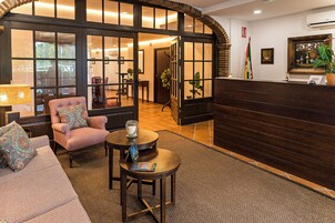 Un área de recepción con un mostrador de madera, una silla acolchada, una mesita con una revista y un sofá con cojines estampados.