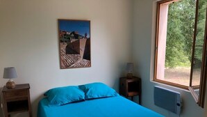 A bedroom with a bed, a nightstand, a lamp, a window, and a wall-mounted heater. - Luberon - Vue Exceptionnelle - Location de Maisonnette 4 Personnes (Ménerbes)