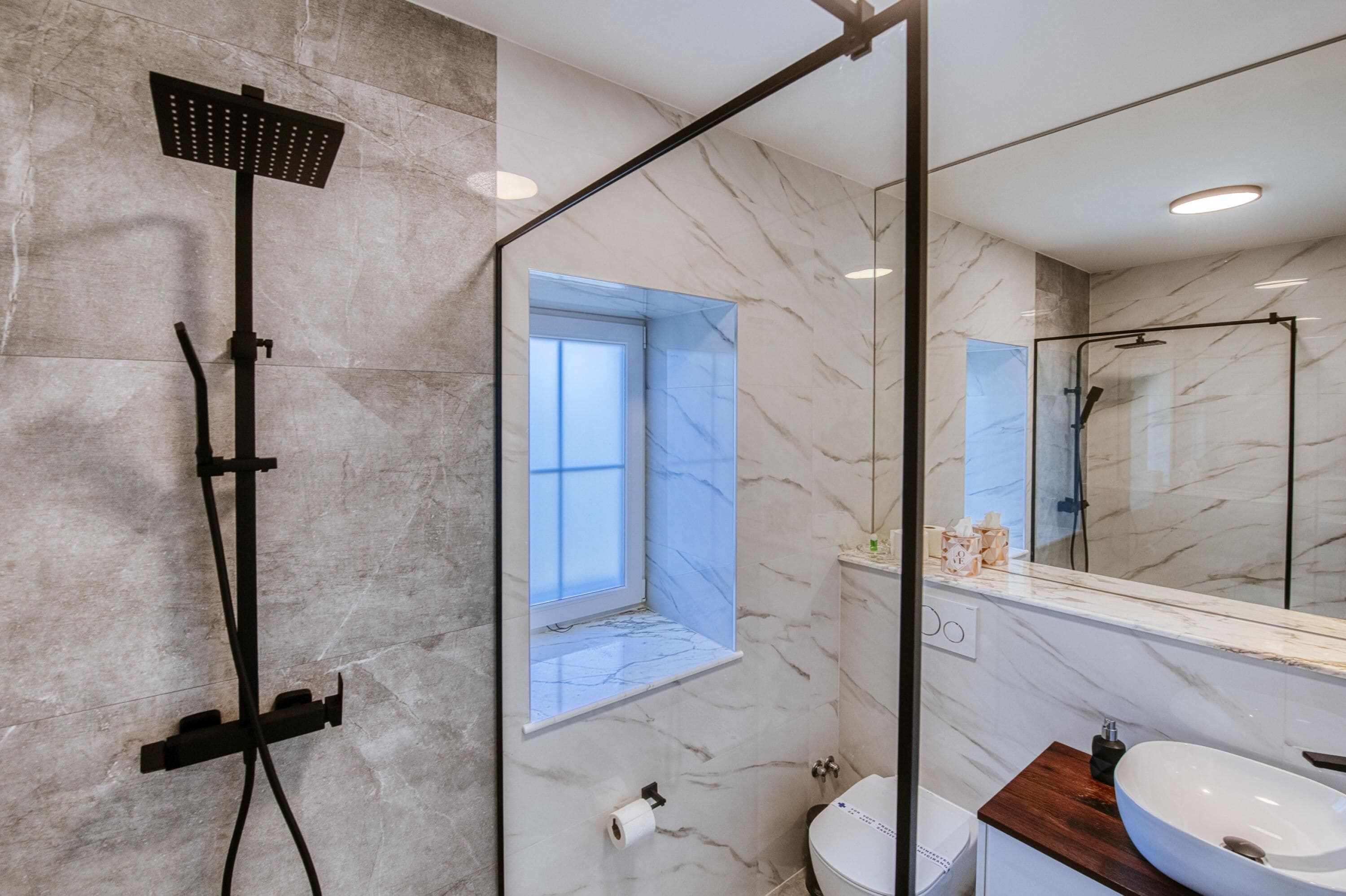 A modern bathroom with a glass-enclosed shower, a marble wall, a window, and a sink with a wooden countertop.