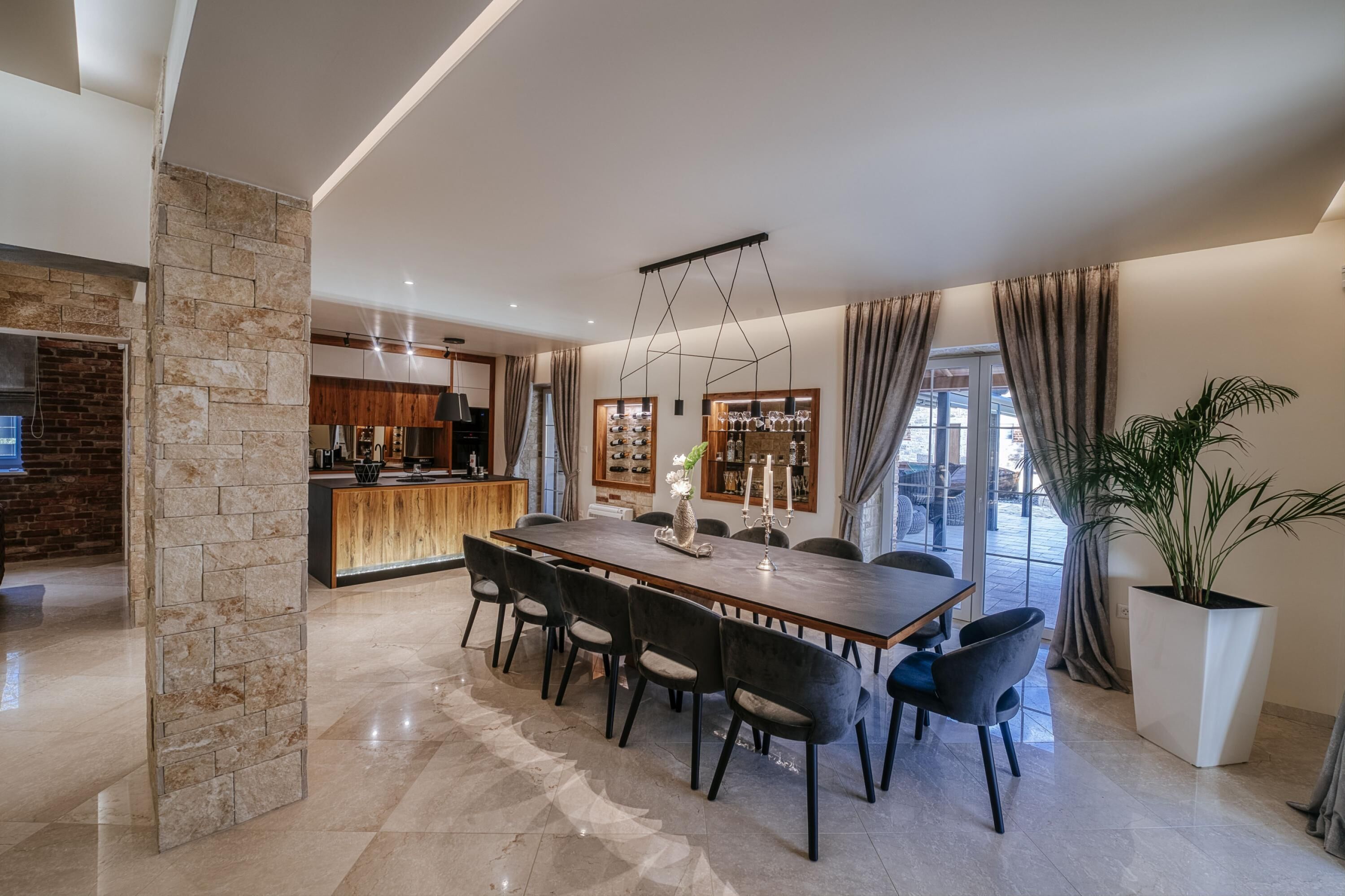 A modern dining area with a long table and chairs, a stone accent wall, a kitchen in the background, and a large potted plant.
