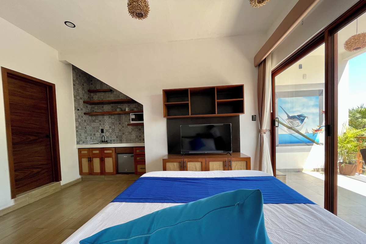 A bedroom with a bed, a television, a kitchenette, and a balcony with a view of the sea.