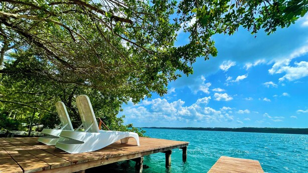 A wooden deck extending over clear blue water, with a white lounge chair under a tree.