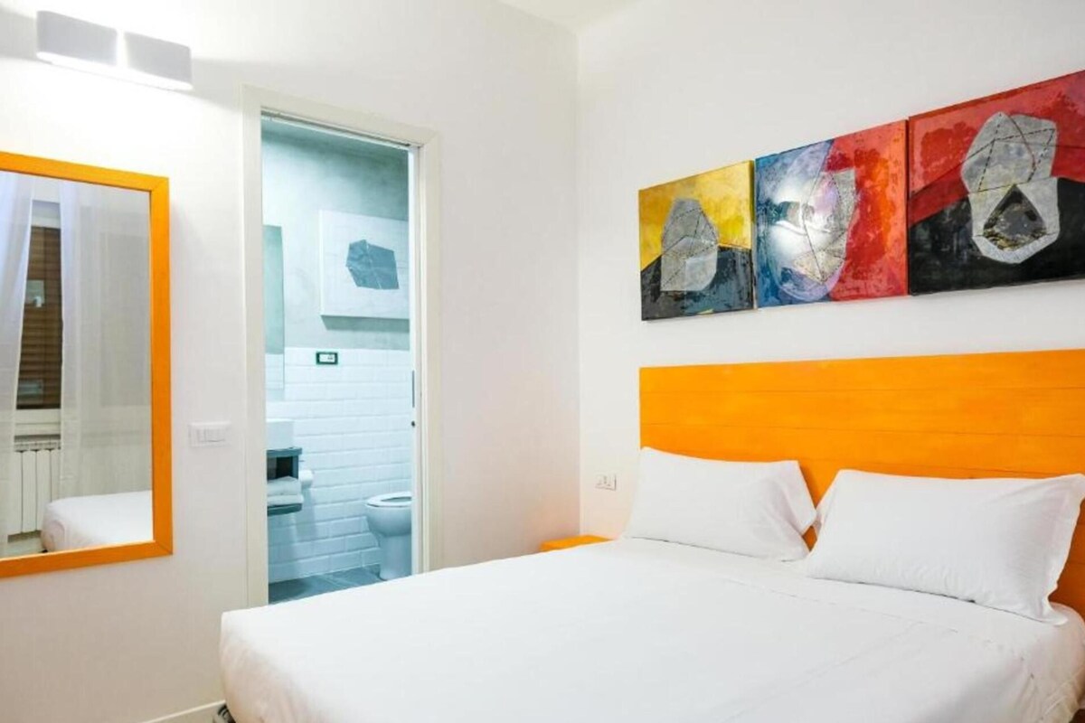 A hotel room with a bed, a mirror, a bathroom visible through an open door, and a colorful abstract painting on the wall.
