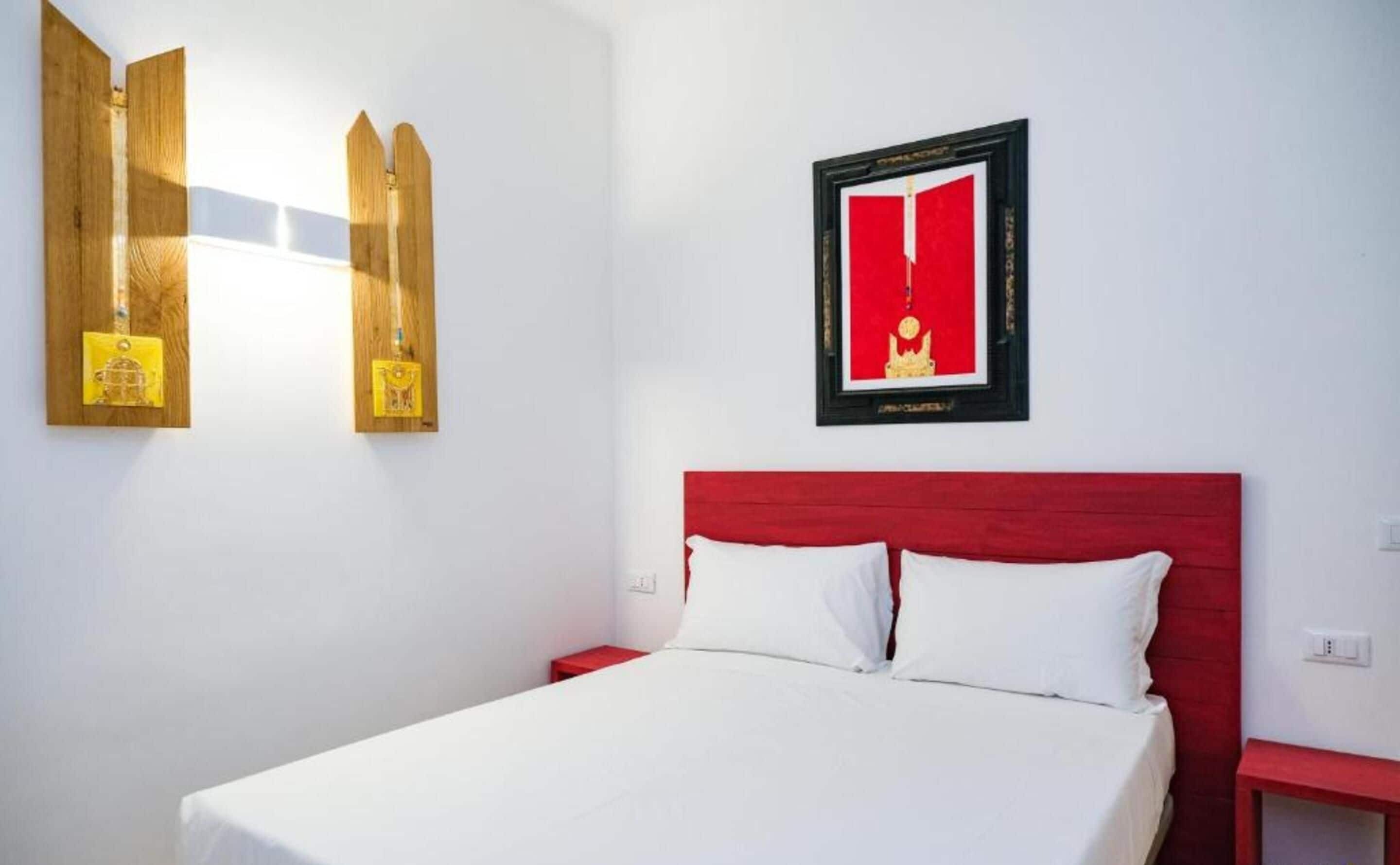 A hotel room with a red headboard, a white bedspread, and two bedside tables. There is a framed picture on the wall and a wooden shelf with decorative items.