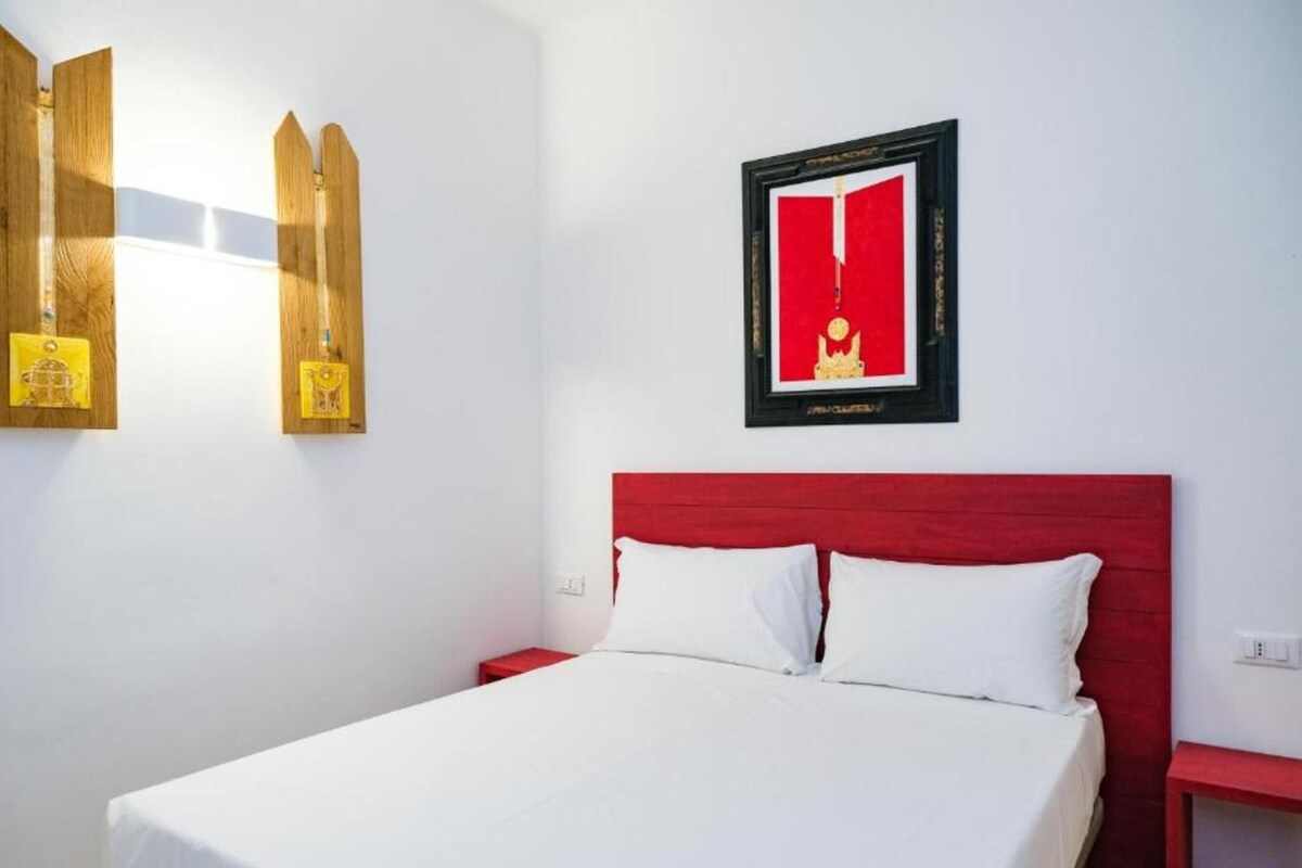Habitación de hotel con cabecera roja, colcha blanca y dos mesitas de noche. Se observa un cuadro en la pared y una estantería de madera con adornos.