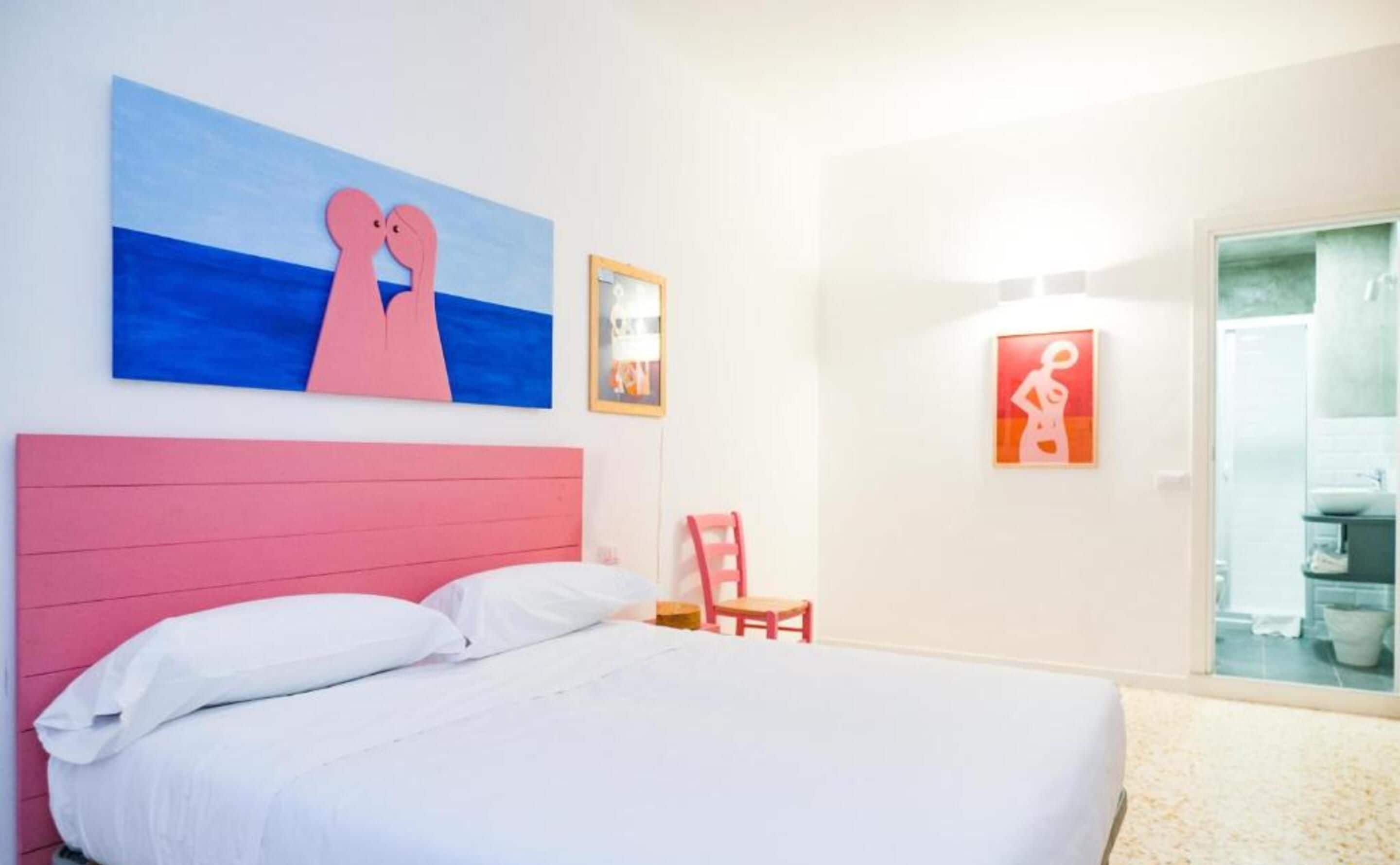 Un dormitorio con cabecera rosa, cama blanca y obras de arte en la pared.