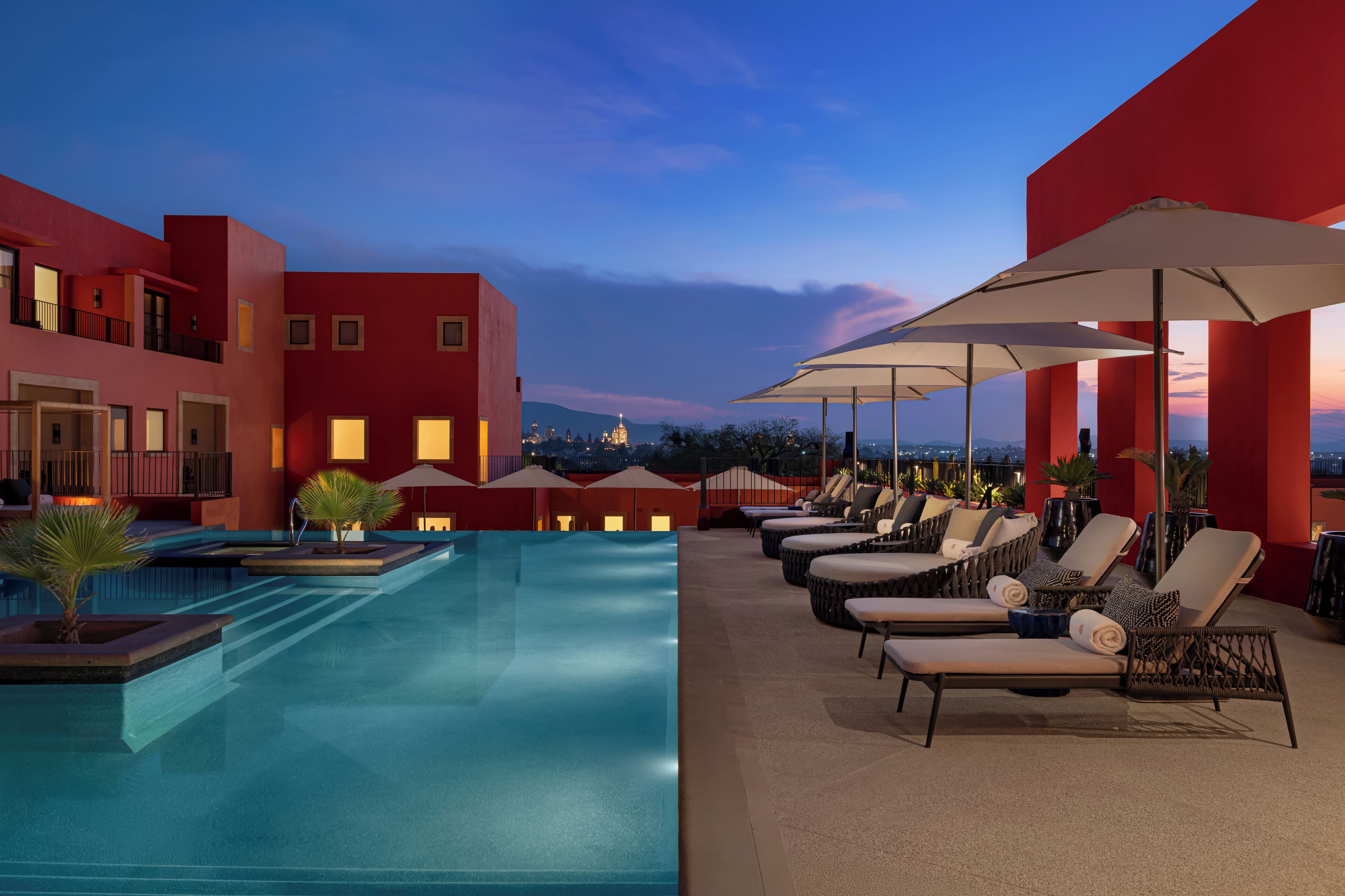 A rooftop pool area with lounge chairs and umbrellas, surrounded by red buildings with illuminated windows.