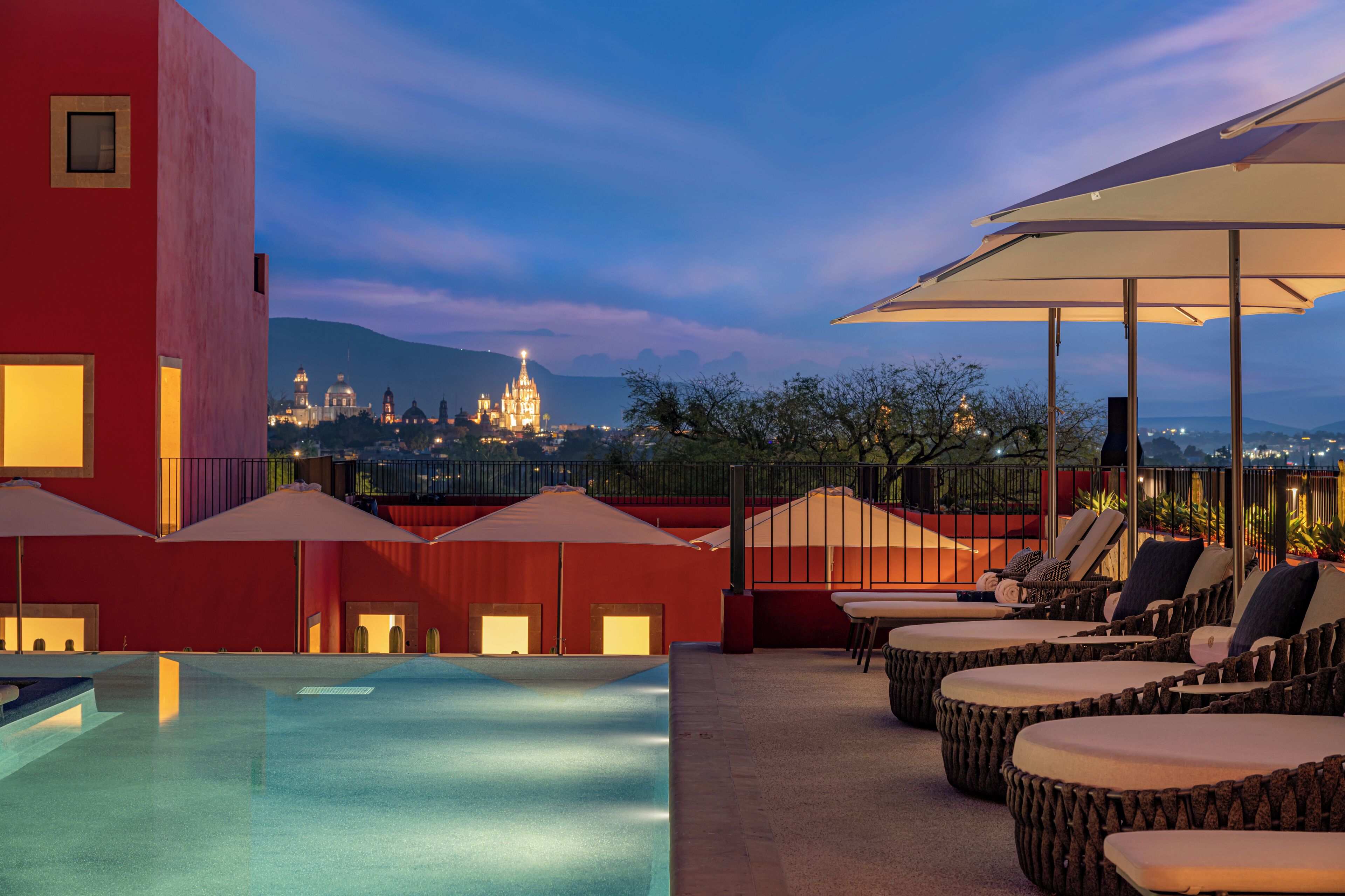 A rooftop pool area with lounge chairs and umbrellas, overlooking a cityscape with illuminated buildings.