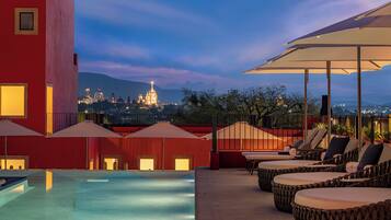 Un área de piscina en la azotea con sillones de descanso y sombrillas, con vistas a un paisaje urbano de edificios iluminados.