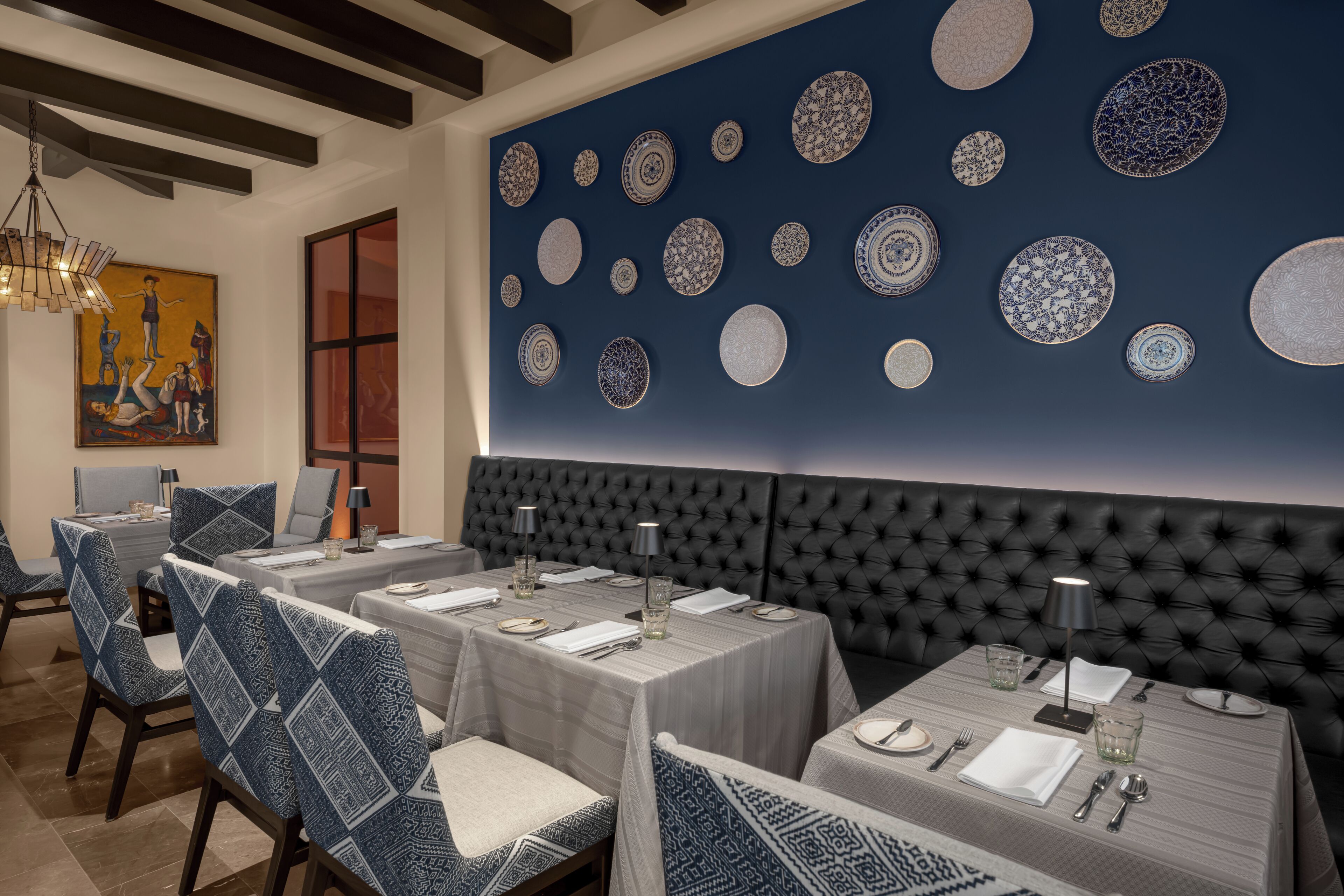 Uma sala de jantar com mesas postas para quatro, uma grande parede com pratos decorativos e uma cabeceira estofada.