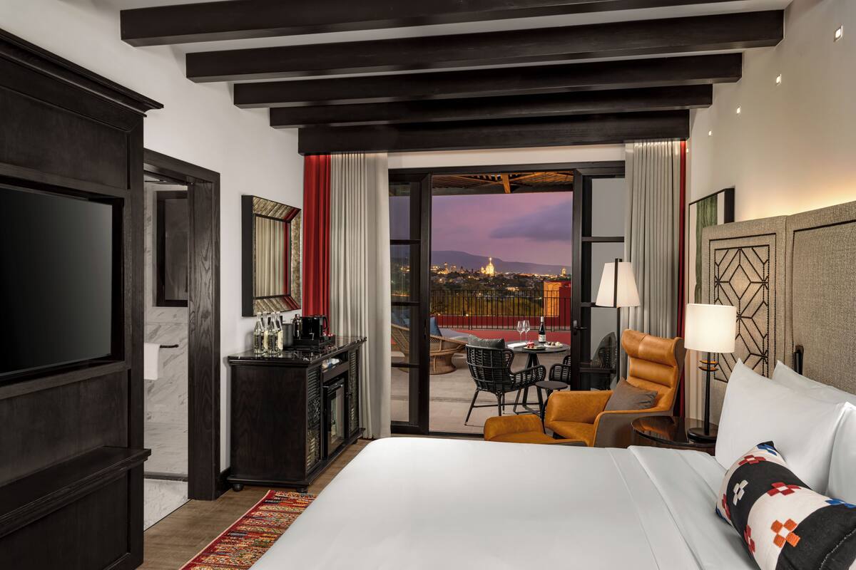 Ein Hotelzimmer mit einem großen Bett, einem Schreibtisch mit Kaffeemaschine, einem Sessel und Blick auf die Stadt durch eine geöffnete Tür.