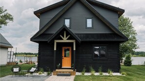 Une maison en bois à deux étages, de couleur noire, avec un toit à pignon, un porche à l’avant et une petite fenêtre au-dessus de la porte.