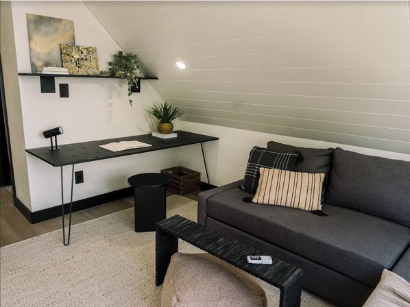 En moderne stue med en mørkegrå sofa, et sort sofabord, et sort skrivebord med en computer og en lille potteplante.