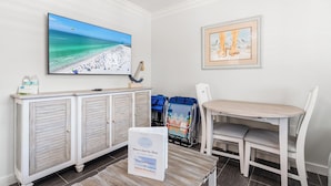 A room with a large TV displaying a beach scene, a framed painting, a white cabinet, and a table with chairs. - One Bed/ One Bath Condo- Step From Siesta Key Beach with Pool On Property! (Siesta Key)