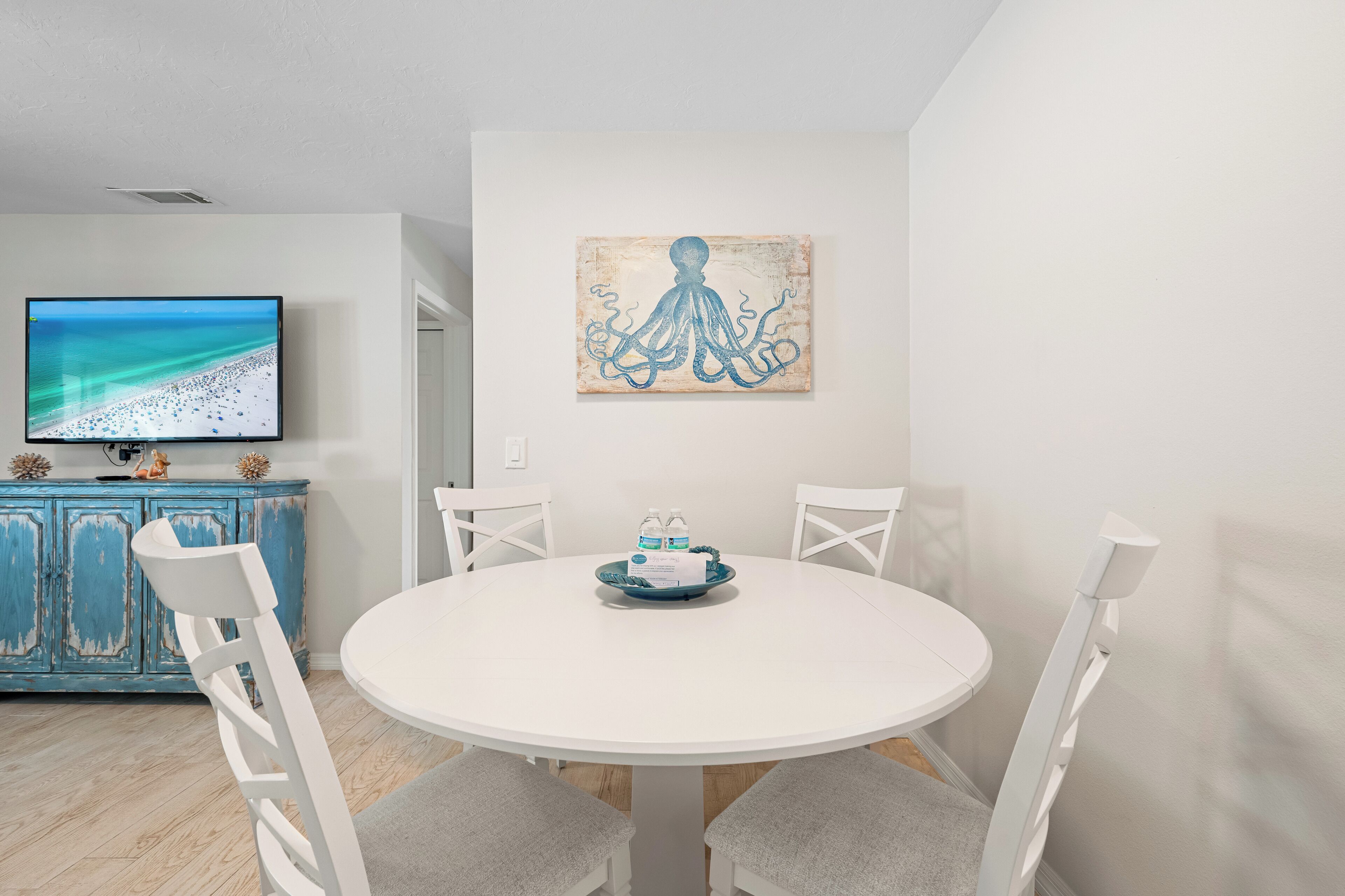 Uma sala de jantar com uma mesa branca redonda, quatro cadeiras brancas, um quadro de um polvo e uma televisão sobre um armário azul.