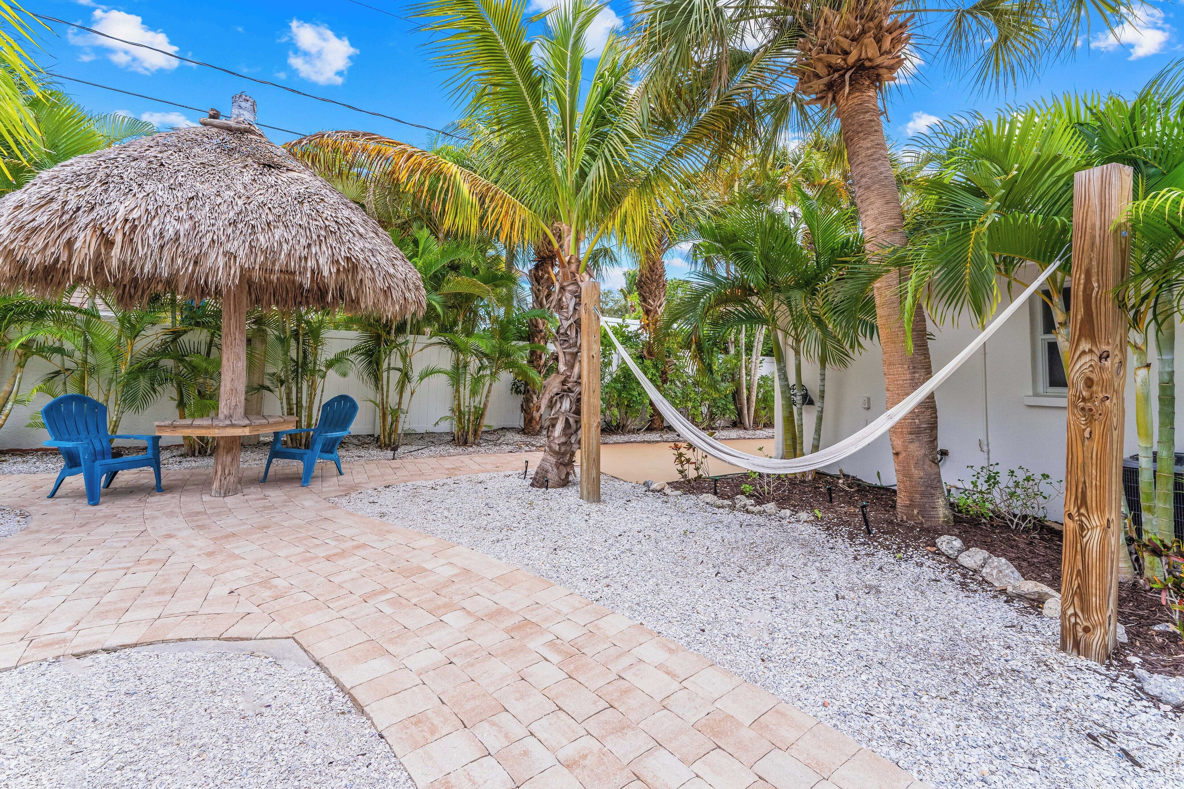 A patio area with a thatched umbrella, two blue chairs, a hammock, and palm trees.