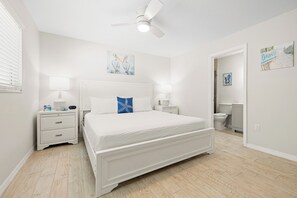 A bedroom with a white bed, two bedside tables, a ceiling fan, and a bathroom visible through an open door.