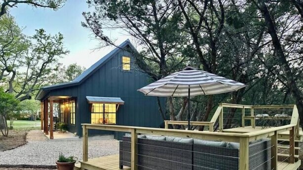 Una terraza de madera con un sofá y una sombrilla, una casa azul con un porche, y un camino de grava.