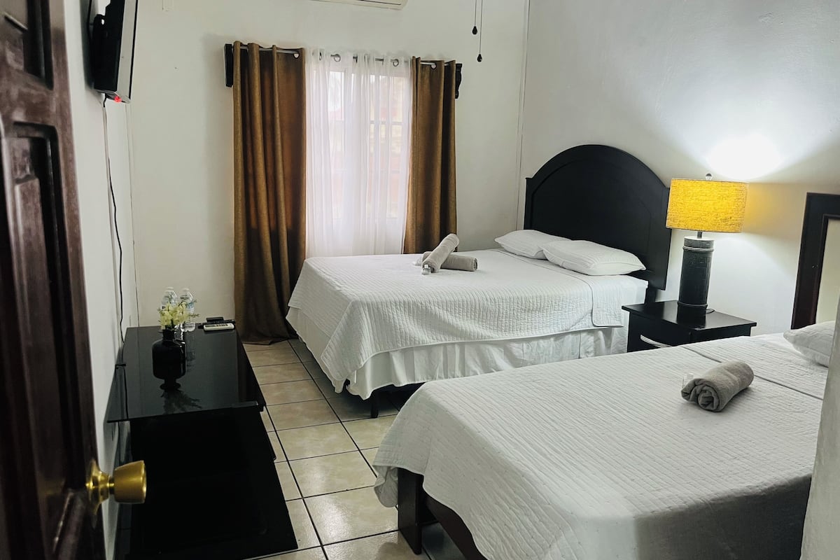 Quarto de hotel com duas camas, mesa de cabeceira com abajur, televisão na parede e janela com cortinas.