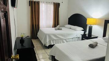 A hotel room with two beds, a bedside table with a lamp, a TV mounted on the wall, and a window with curtains.