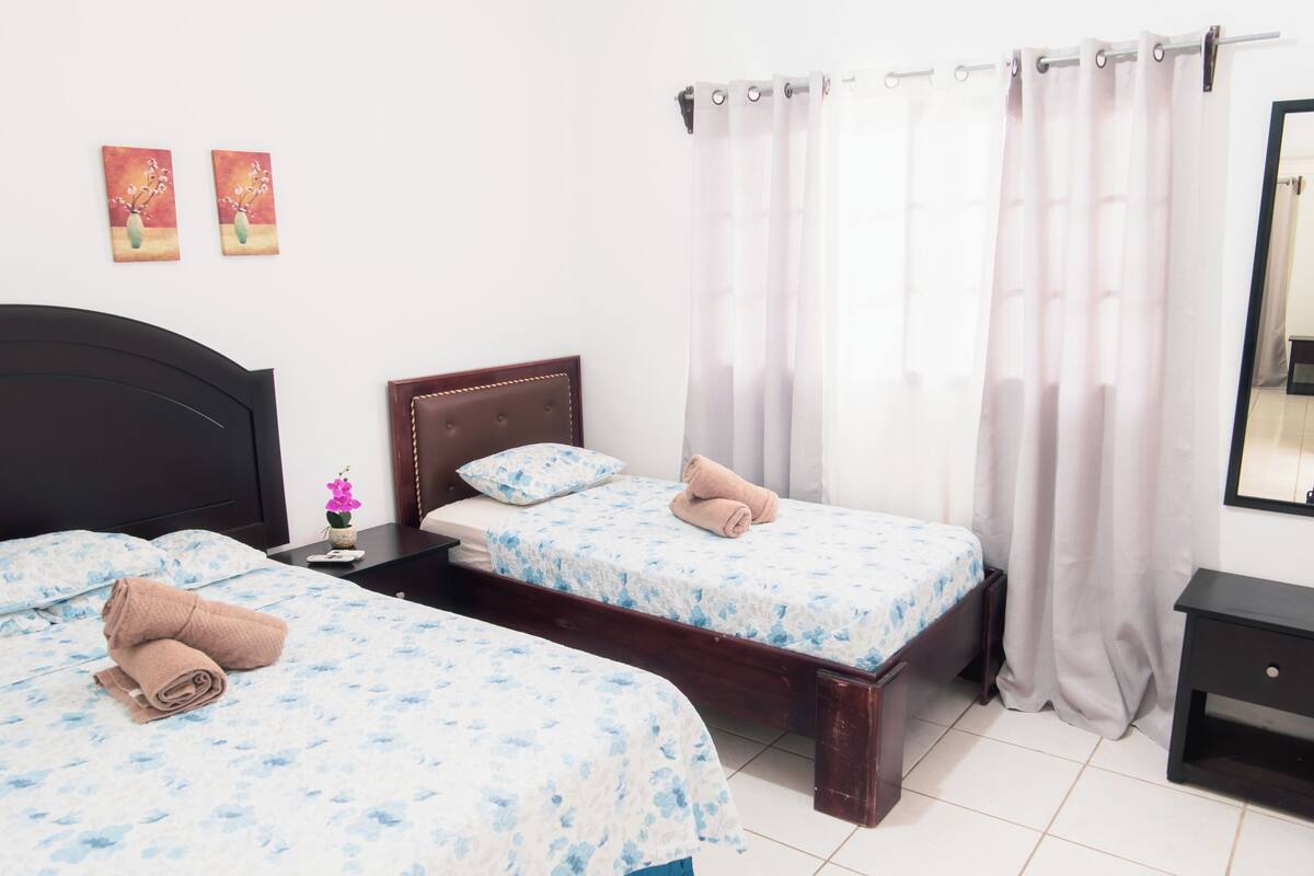 Quarto de hotel com duas camas de solteiro, cabeceira de madeira, uma mesa pequena com um vaso de flores e um espelho na parede.