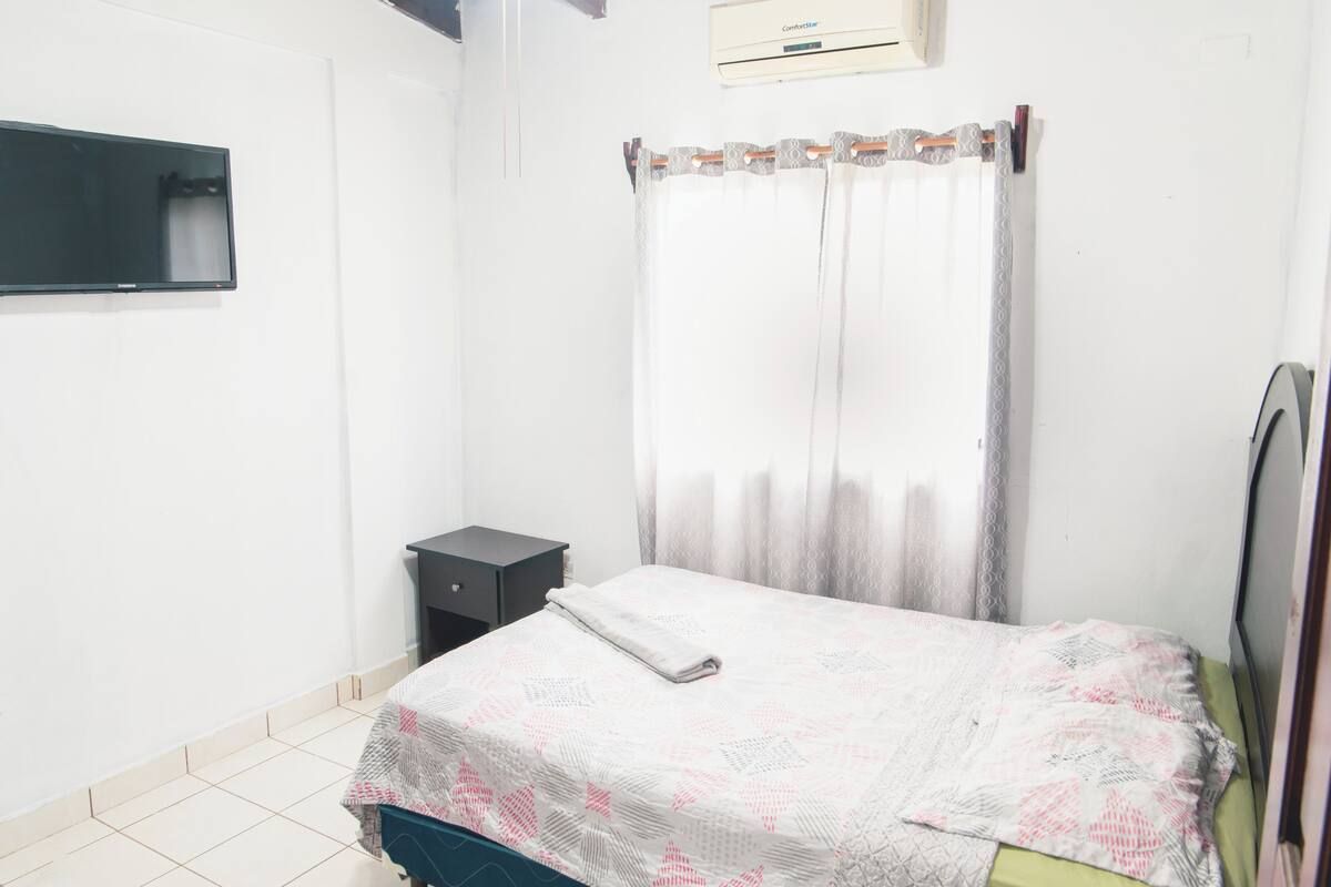 Quarto com cama, mesa de cabeceira pequena, televisão na parede e janela com cortinas.