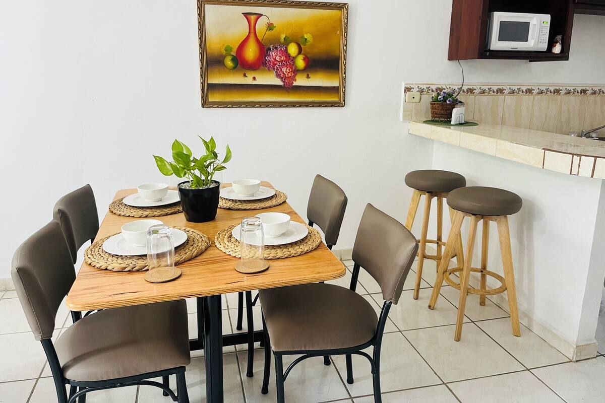 Uma sala de jantar com uma mesa de madeira posta para quatro pessoas, rodeada por cadeiras, e uma cozinha com micro-ondas e banquetas.
