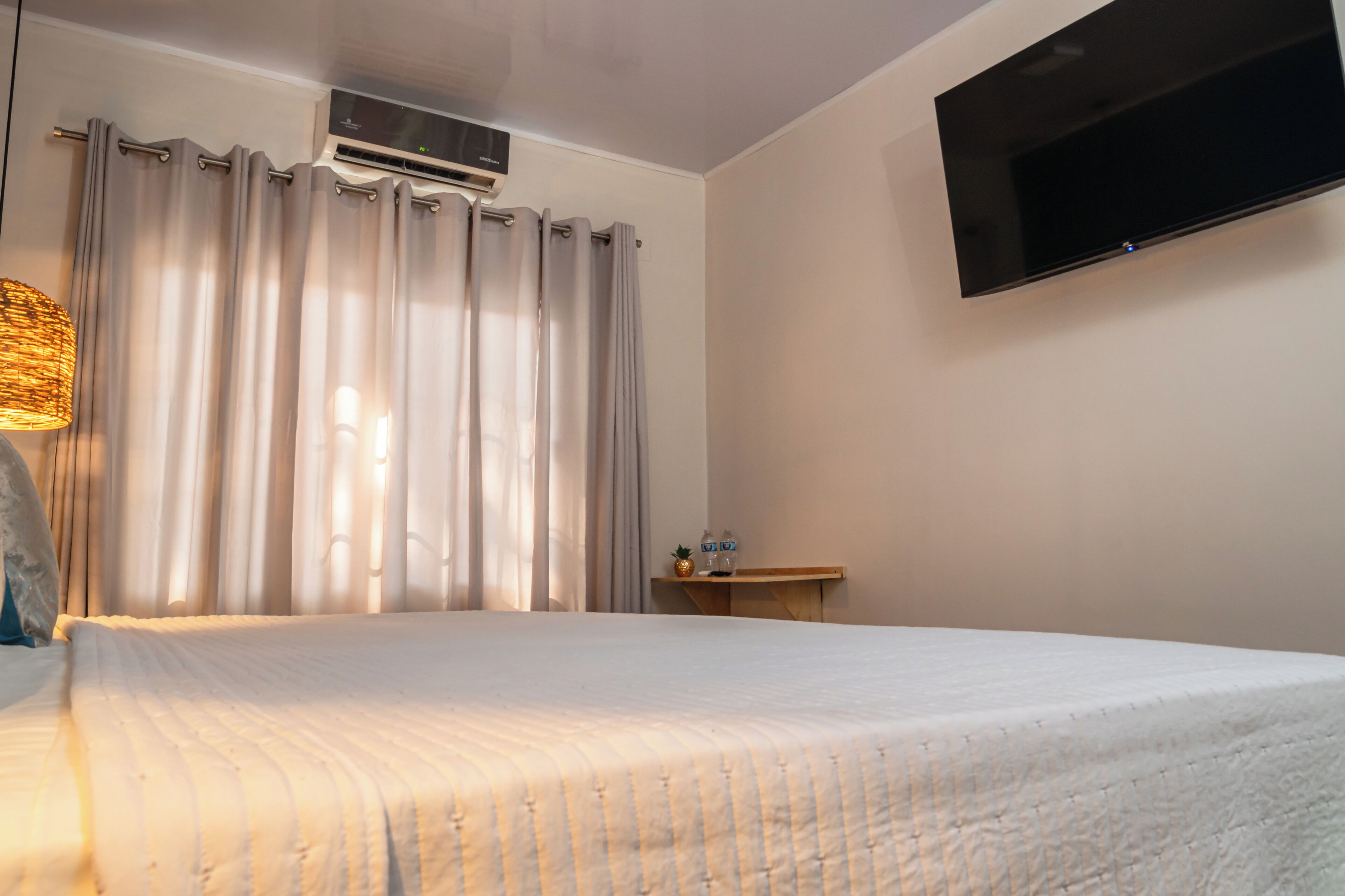 Habitación de hotel con cama, cortinas, aire acondicionado, televisor montado en la pared y una mesita con una botella y un vaso.