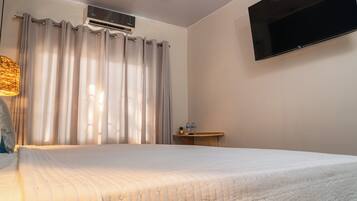 Habitación de hotel con cama, cortinas, aire acondicionado, televisor montado en la pared y una mesita con una botella y un vaso.