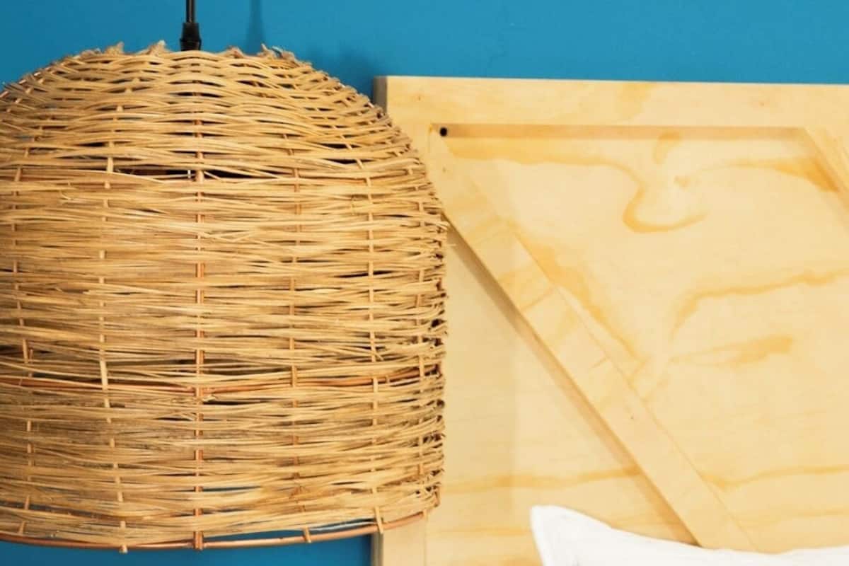 Luminária pendente de vime suspensa por um cordão preto, cabeceira de madeira com travesseiro branco e parede azul.