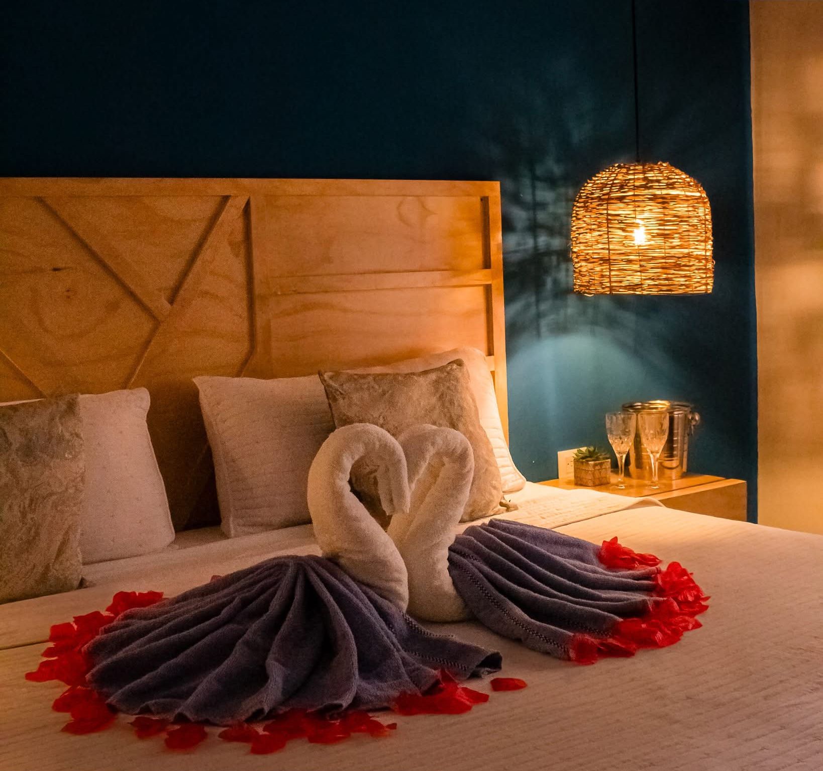Una habitación de hotel con cama, almohadas, cabecero, mesita de noche con vasos y una planta pequeña, y una lámpara colgante.
