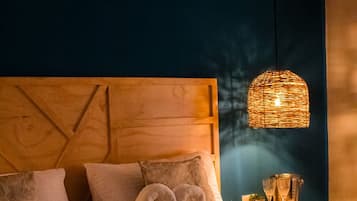 Una habitación de hotel con cama, almohadas, cabecero, mesita de noche con vasos y una planta pequeña, y una lámpara colgante.