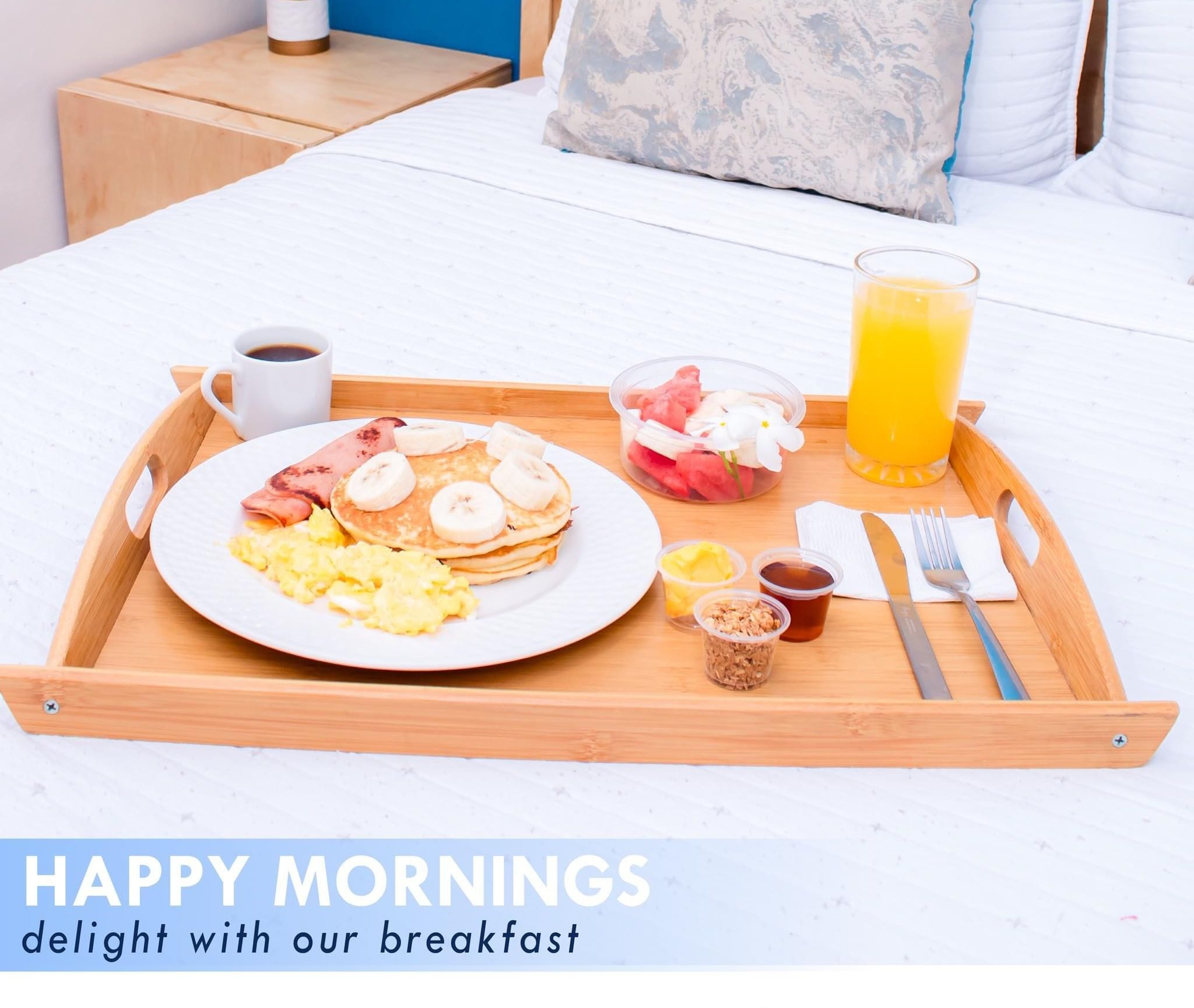 Bandeja de desayuno con panqueques, huevos, fruta y jugo sobre una cama en una habitación de hotel.