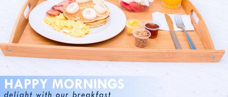 Bandeja de desayuno con panqueques, huevos, fruta y jugo sobre una cama en una habitación de hotel.